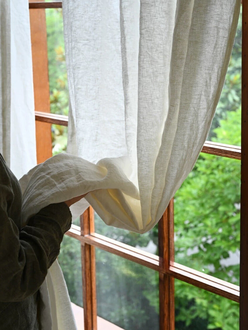 Sheer white linen curtain hanging by wooden window with hand touching fabric