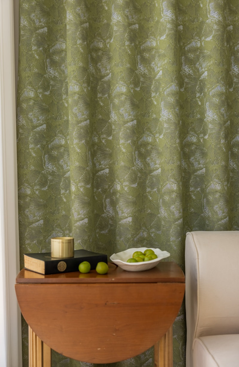 Green jacquard curtains with ginkgo leaf pattern hanging behind wooden table with books and bowl of green grapes in cozy living room setting