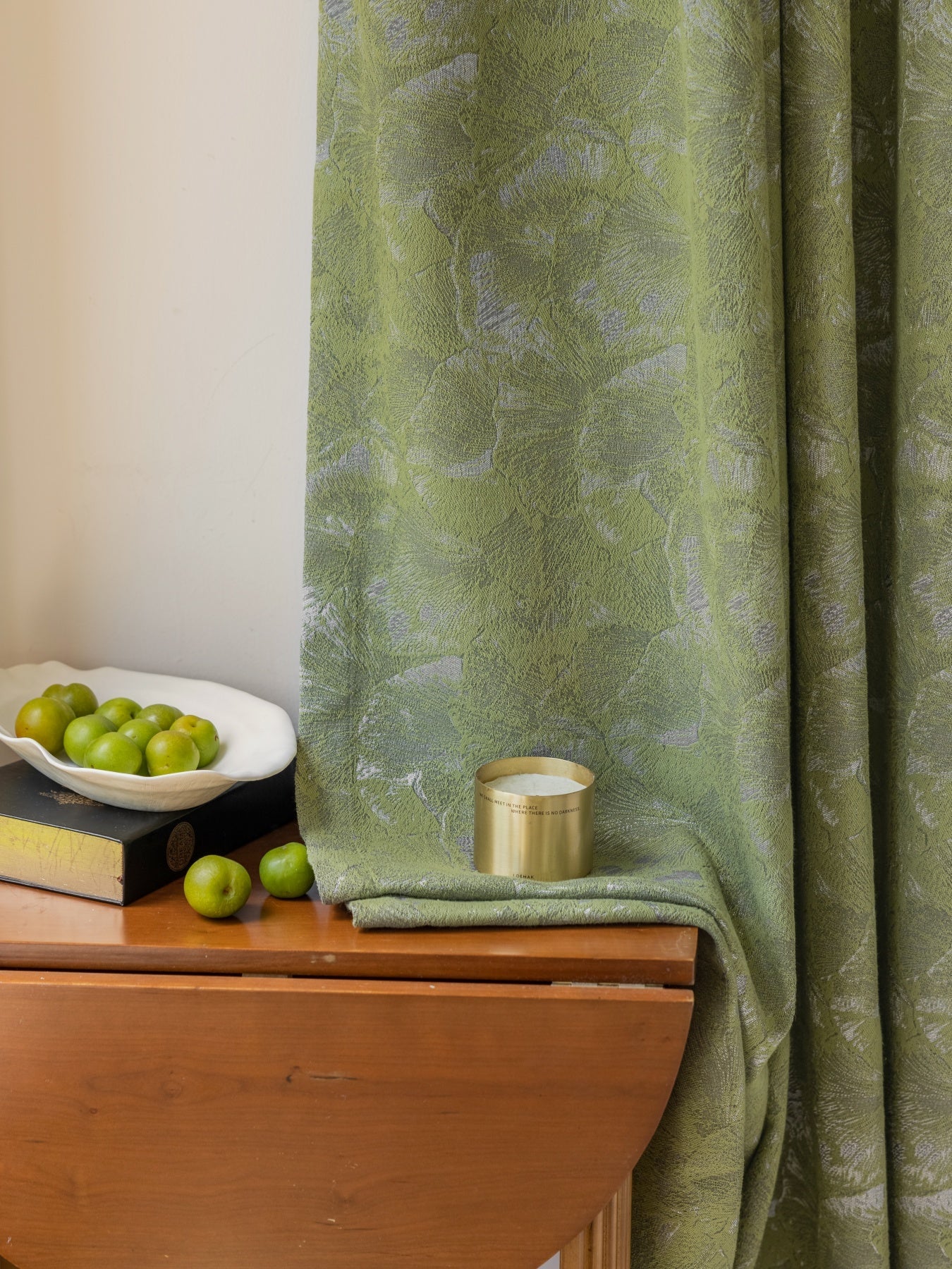 Green jacquard curtain with ginkgo leaf pattern hanging beside a wooden table with green apples and a candle.