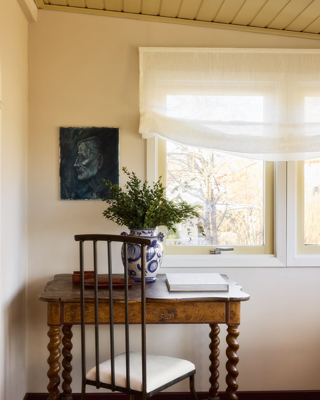 White soft fold sheer Roman shade over window in cozy room with wooden desk and chair