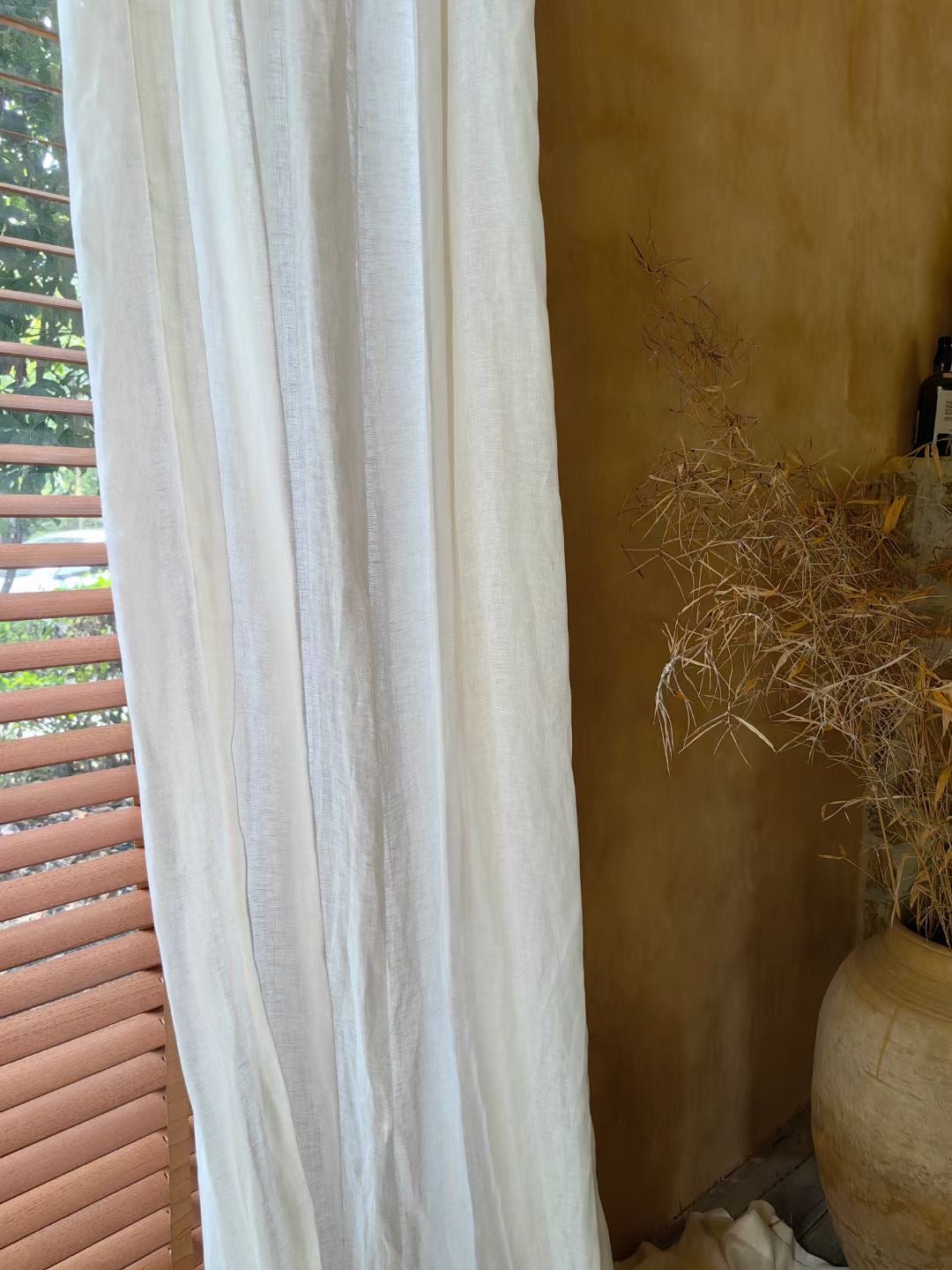 white sheer linen curtain hanging by window with wooden blinds and decorative dried plant in rustic room