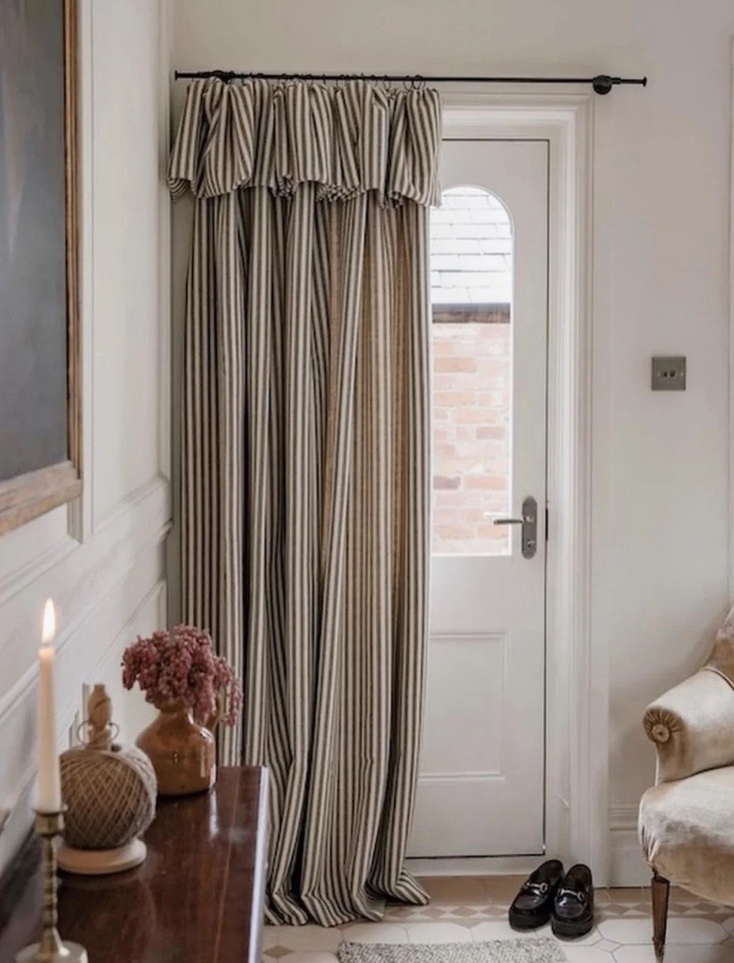 Beige linen striped curtains with ruffled top hanging by a white door in a cozy, softly lit room.