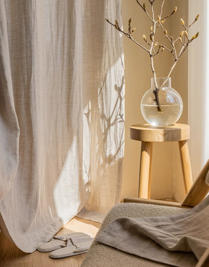 Elegant sheer linen curtain in natural tone with sunlight filtering through, accompanied by a wooden stool and vase with branches, highlighting quality French linen drapery