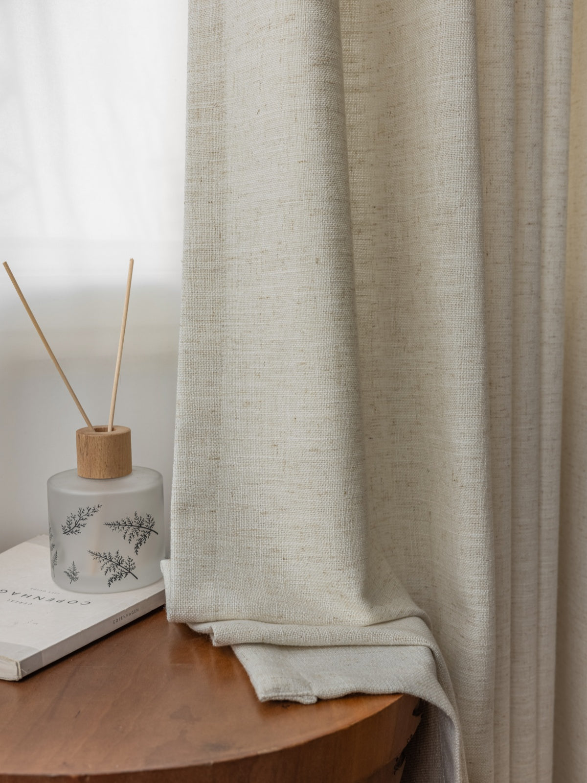 Luxurious white French linen blend drape pleated, enhancing room ambiance with a side table featuring a fragrance diffuser and botanical book