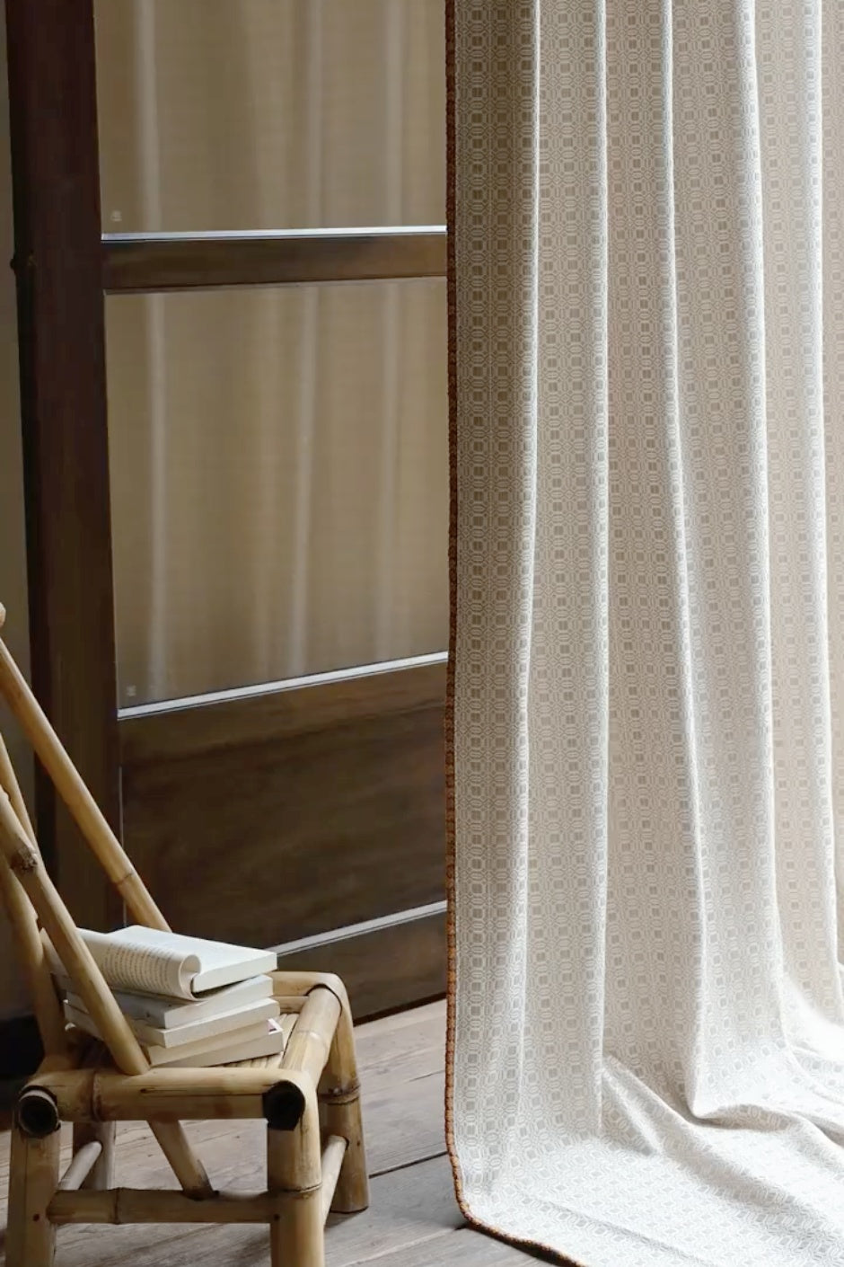 Vintage curtains with subtle pattern and brown trim hanging beside a wooden window and wicker chair with stacked books in a cozy room setting
