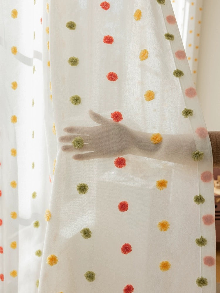 Close-up of sheer curtain with colorful rainbow pom-poms in a kid's room, hand touching the whimsical, eco-friendly fabric
