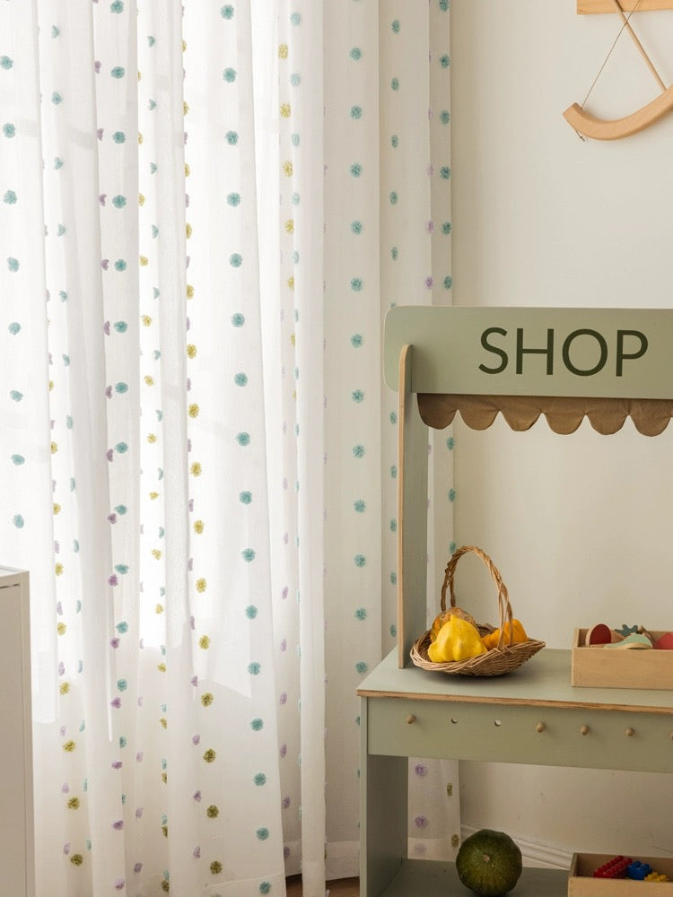 Colorful pom-pom adorned sheer curtains in a child's room with a playful 'shop' stand and a basket of bananas