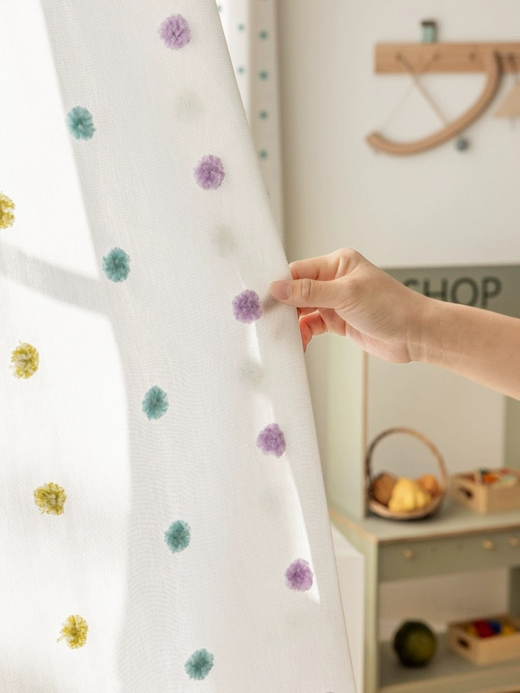 Close-up of a hand brushing colorful pom-pom embellishments on white sheer curtains in a child's room, perfect for adding playful charm and whimsy