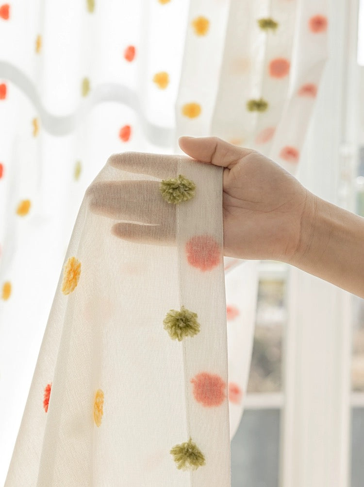 Hand holding colorful candy-themed sheer curtain with red, orange, yellow, and green pom-poms, ideal for kids' rooms, enhancing a playful and charming interior atmosphere