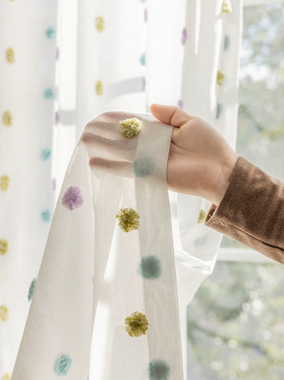 Colorful candy sheer curtains with rainbow pom-poms in a kids' room, hand demonstrating the soft, plushy texture against a sunlit window