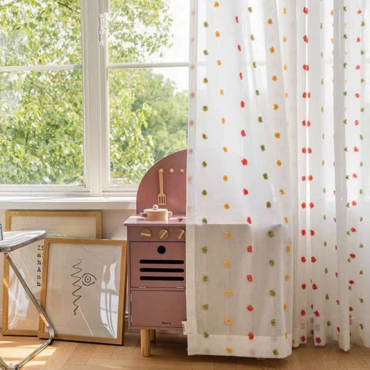 Colorful candy sheer curtains with pom-poms in a child's room beside a pink cabinet and framed artworks, enhancing playful room decor