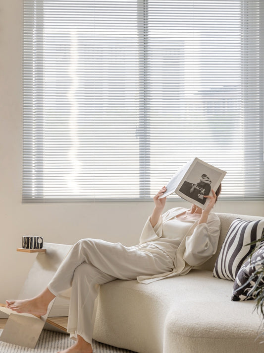 Relaxed individual reading a book on a sofa with EaseEase cordless vinyl mini blinds filtering sunlight in the background