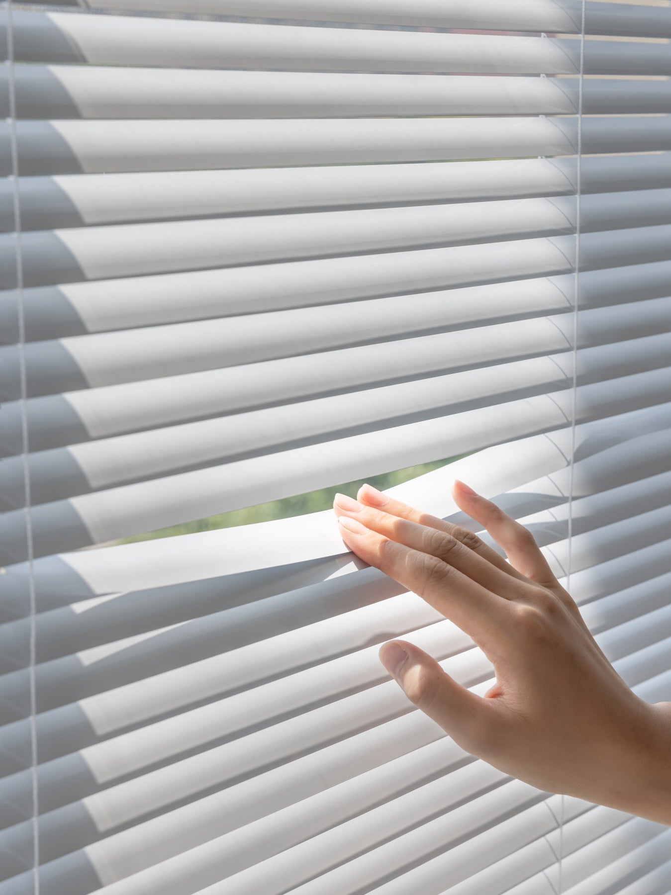 Close-up of a hand adjusting white cordless vinyl mini blinds, letting sunlight filter through, showing ease of use and stylish simplicity