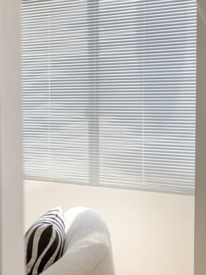 Stylish white vinyl mini blinds in a sunlit room with a cozy armchair, demonstrating easy installation and cleanability
