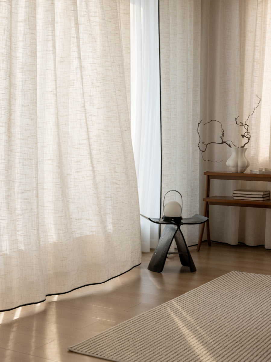 High-quality neutral linen drapes in a stylish living room with natural light, complemented by a minimalist stool, decorative branch, and textured rug