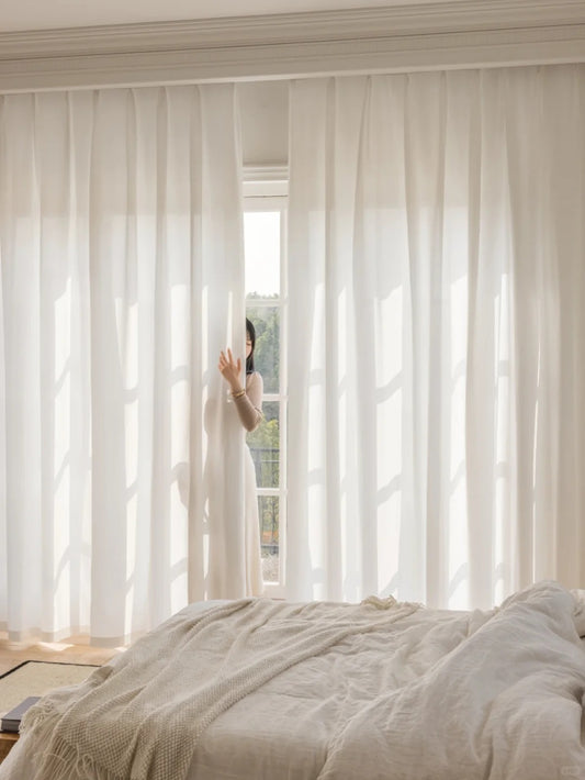Serene bedroom with white sheer curtains providing privacy and soft light penetration, hand slightly pulling curtain aside