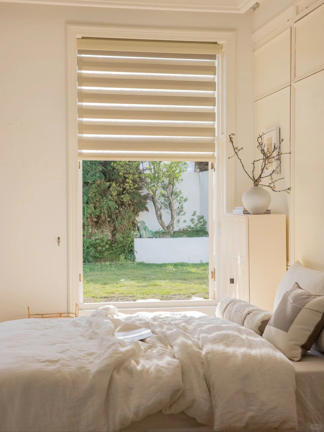 Bedroom window with beige cordless zebra blinds partially open, letting natural light and outdoor greenery in, showcasing modern elegant waterproof shades.