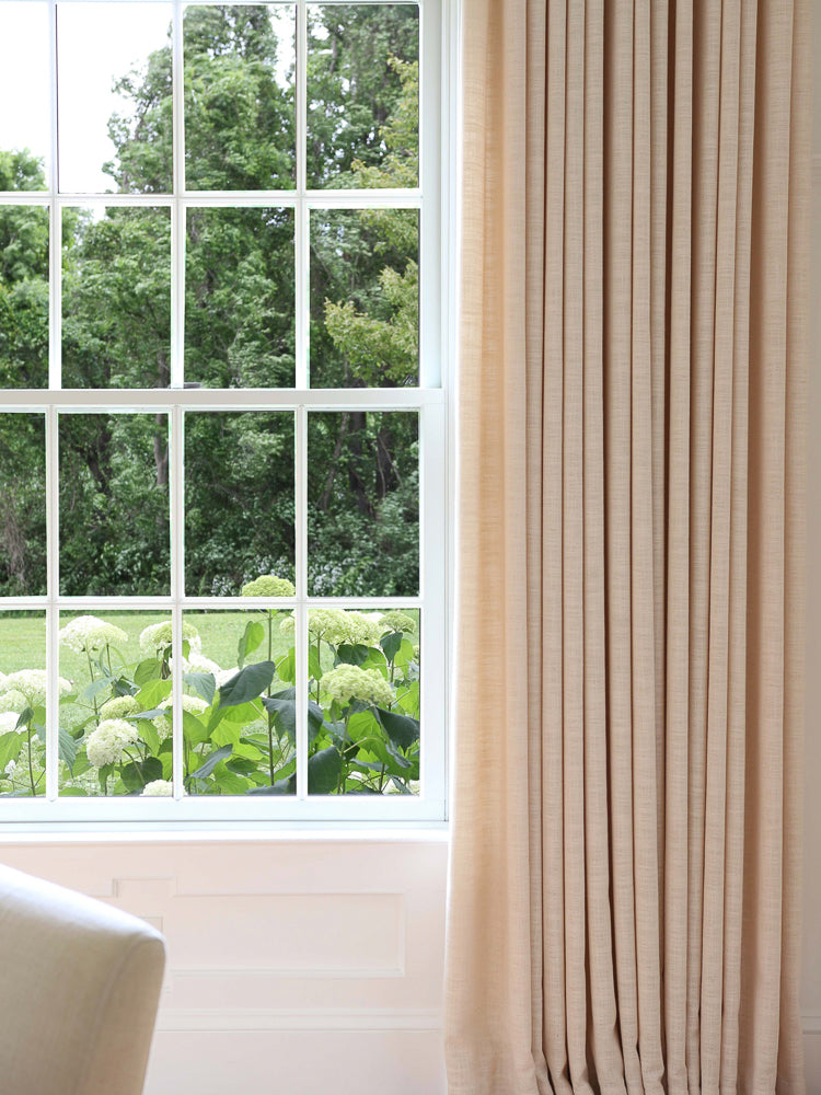 Hunter style linen blackout curtains in a neutral hue, elegantly hanging beside a window with a garden view.