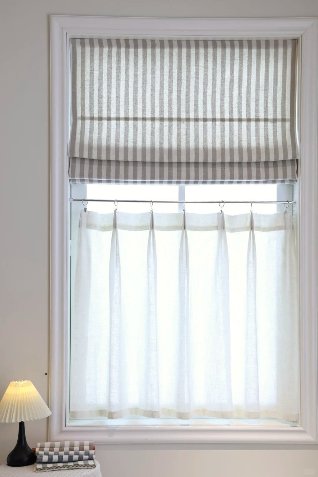 Striped Roman Shades in beige with light filtering linen, cordless lift, installed on a white window frame with sheer curtains underneath and a lamp on the side