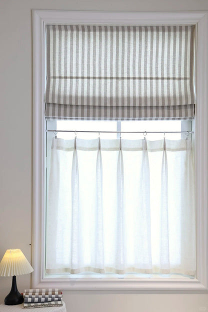 Striped Roman Shades in beige with light filtering linen, cordless lift, installed on a white window frame with sheer curtains underneath and a lamp on the side
