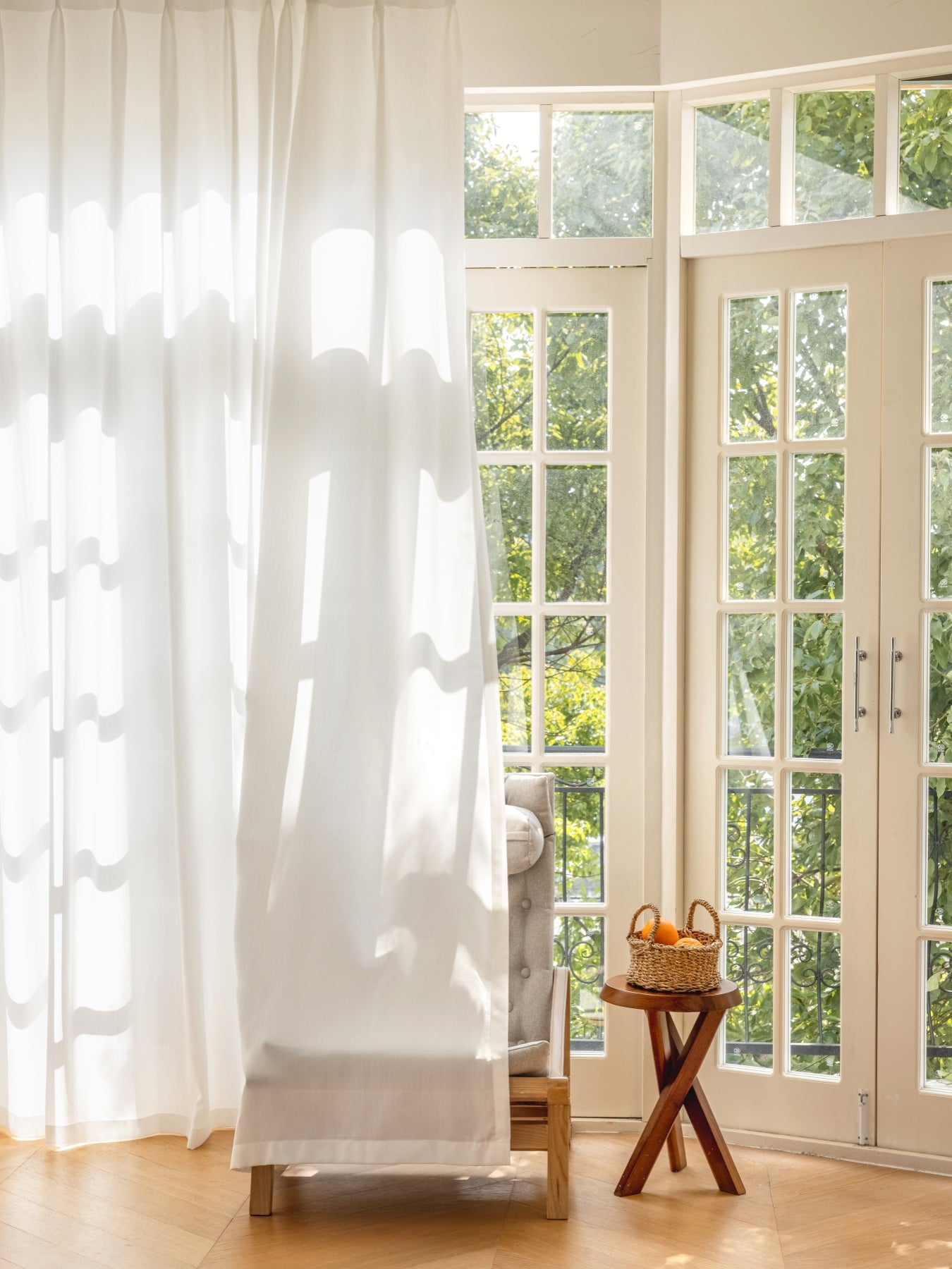 Elegant white sheer curtain filtering sunlight in a room with French doors and a wooden stool with towels, enhancing privacy and soft ambiance