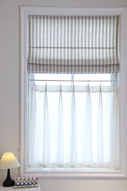 Striped Roman shades with cordless lift in light filtering fabric on window with sheer curtain and bedside lamp
