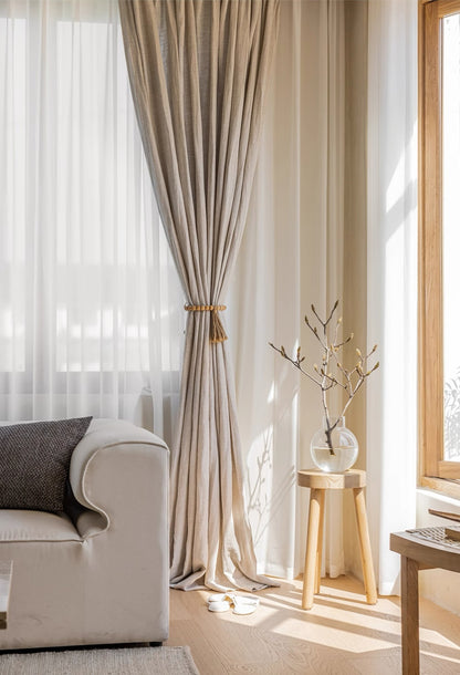 Natural linen curtains sourced from France elegantly draped in a sunlit room, complemented by a cozy armchair and stylish wooden accents
