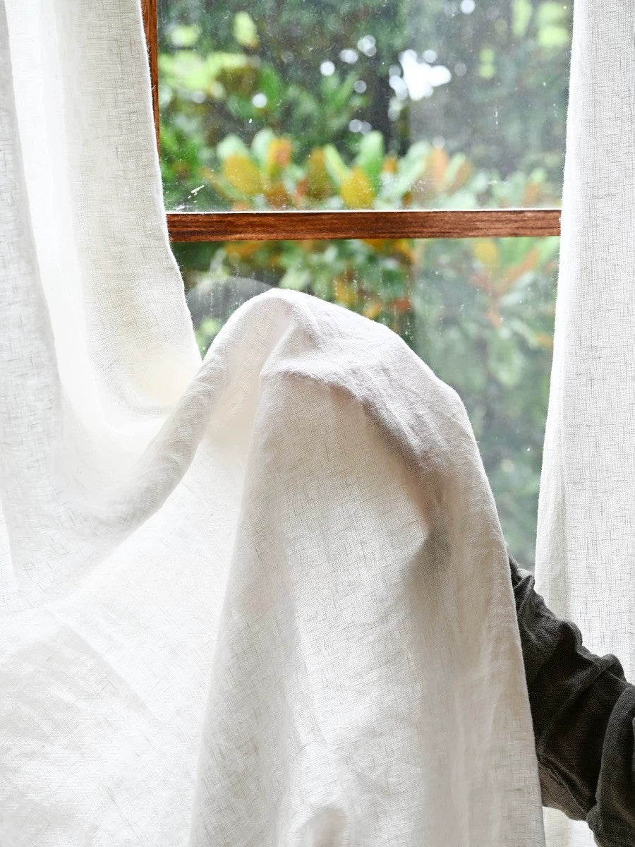 sheer white linen curtain draping by window with blurred outdoor greenery background