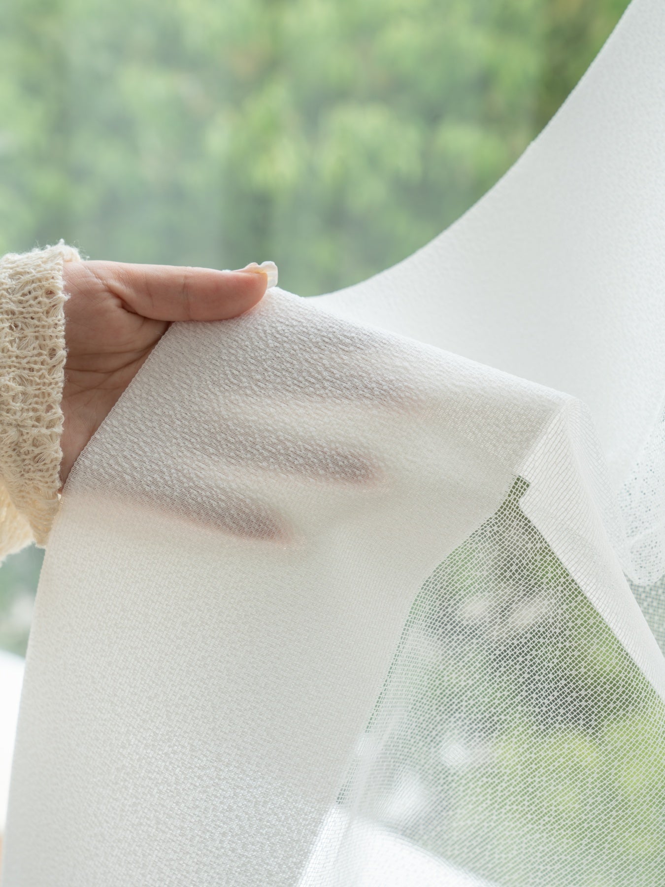 Close-up of hand holding textured semi-transparent curtain by window, showcasing stain-resistant and moisture-resistant features for dreamy elegance.
