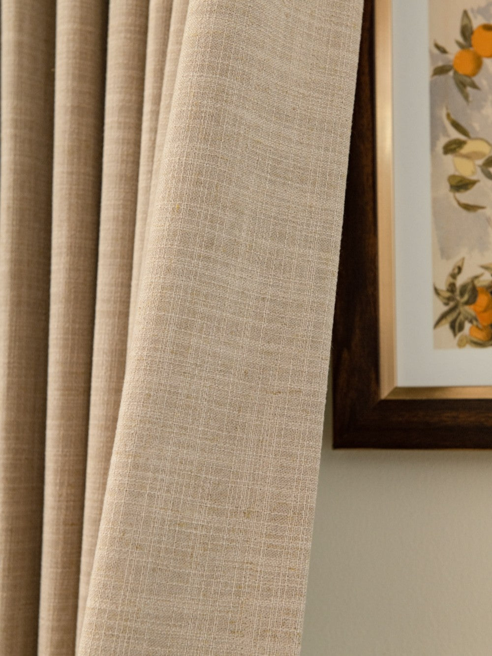 Close-up view of beige Eco-friendly linen blend pleated curtain, featuring a detailed weave texture, next to a framed botanical painting