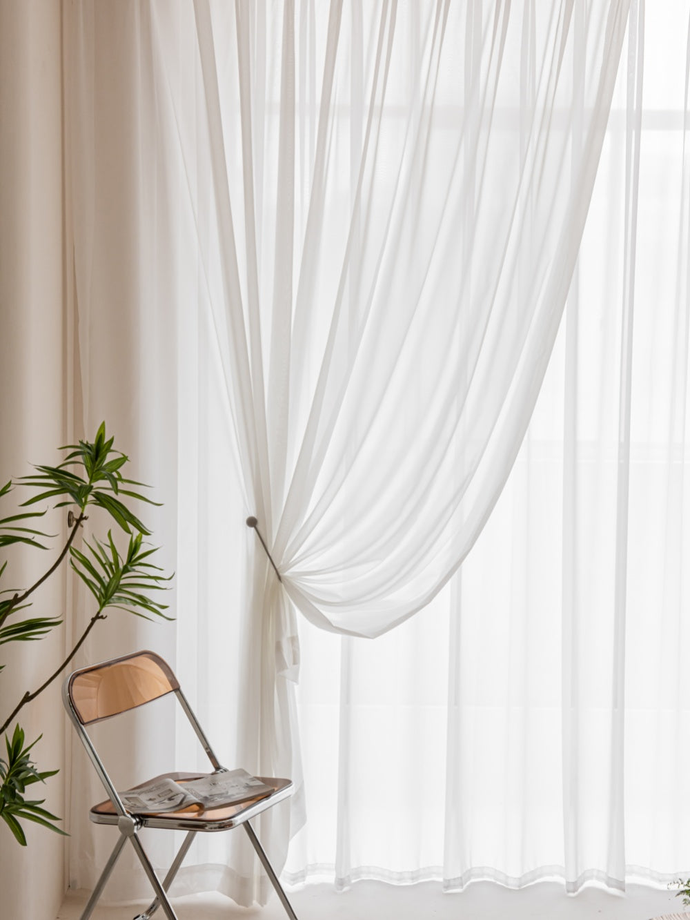 Elegant white gauze sheer curtain tied to the side in a bright room with a wooden chair holding books and a potted plant, embodying serene home decor