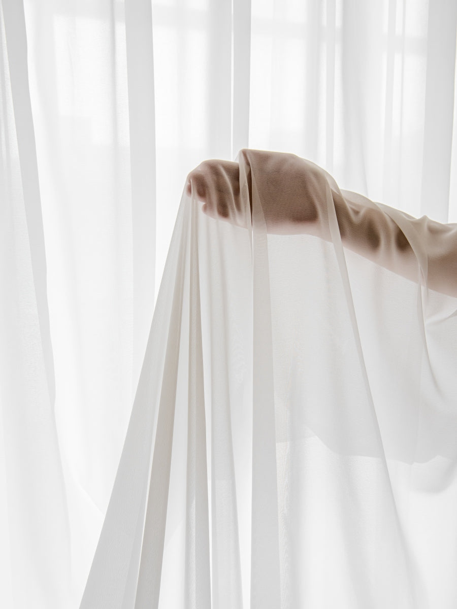 Close-up of a hand drawing aside a thin white sheer curtain, illustrating the light, airy quality and semi-transparency of the fabric