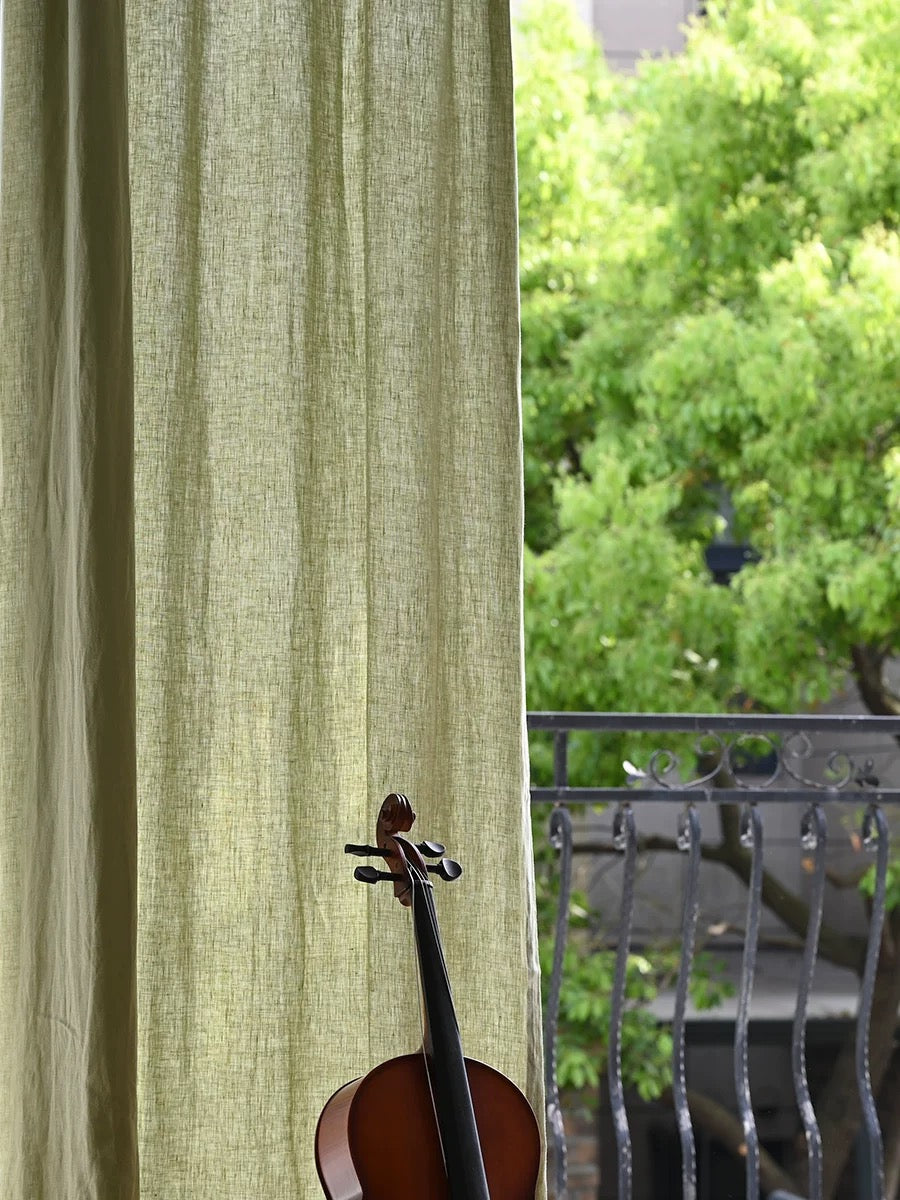 Sage green linen curtains with natural texture softly draping by a window overlooking greenery and a balcony railing.