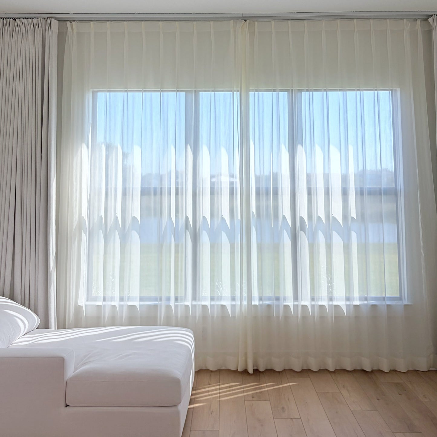 Sheer curtains in cream and white with radiant shimmer, filtering natural light in a bright sunroom with wooden floor and white sofa