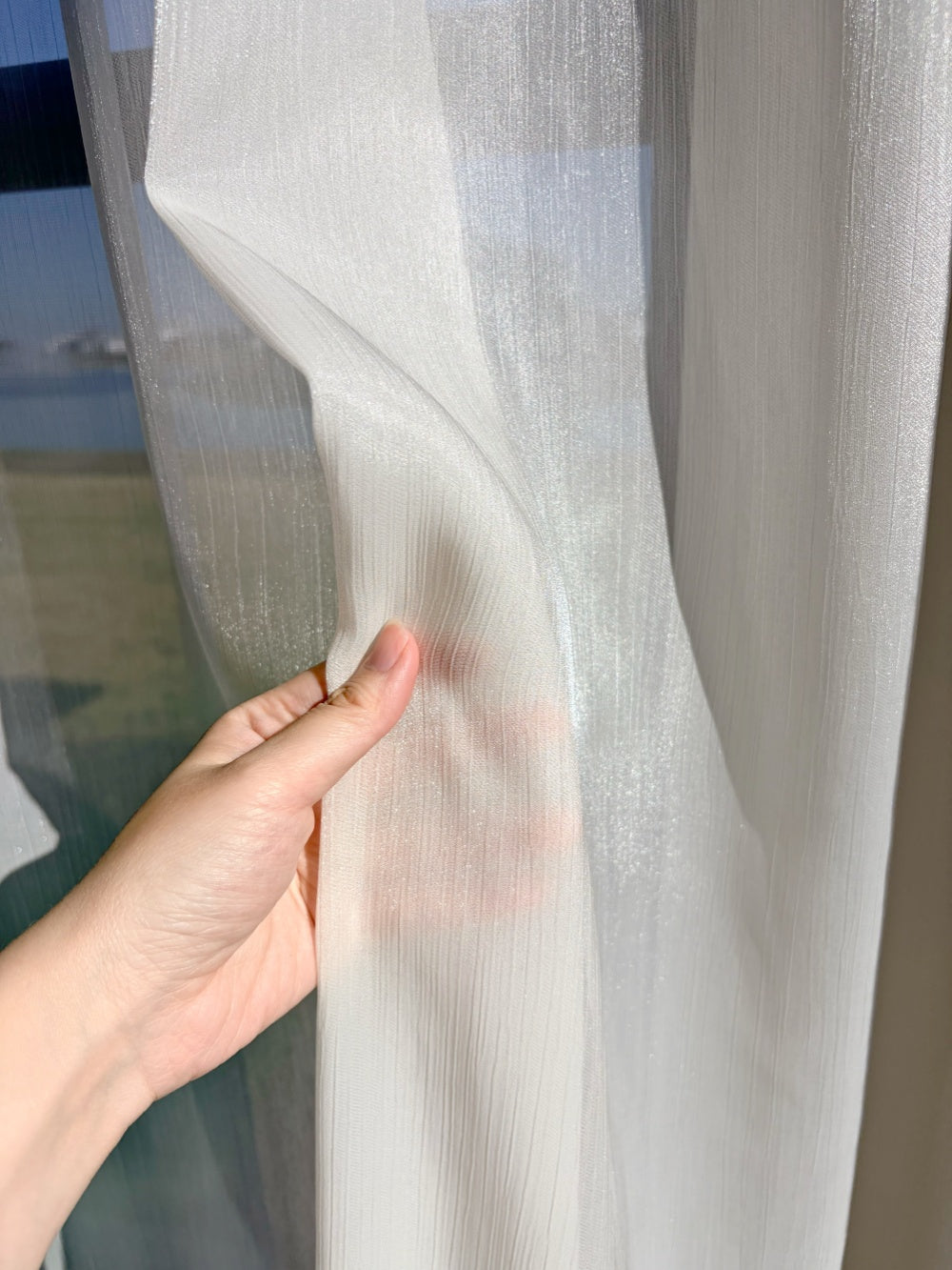 Hand holding cream sheer curtain with sunlight filtering through in sunroom environment