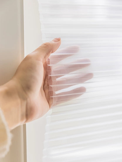 Close-up of a hand adjusting a semi-transparent honeycomb shade with natural light filtering through, demonstrating the product's deliate texture and design quality.