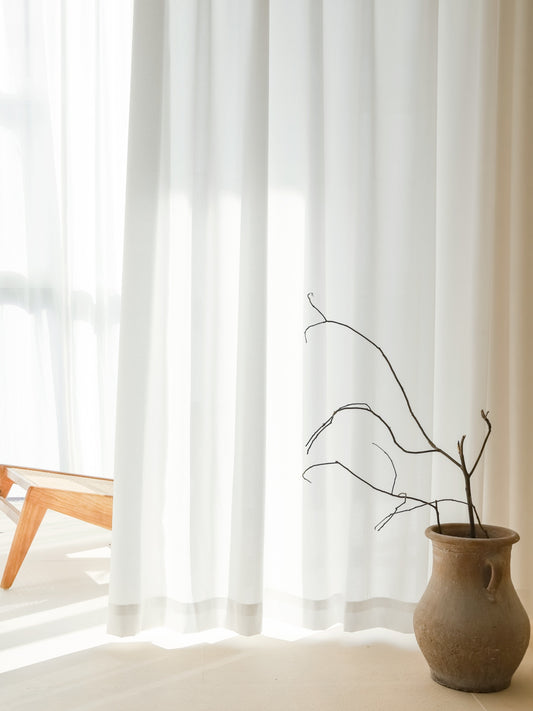Elegant sheer white sun-blocking curtain in a serene room setup with natural light filtering through, complemented by a rustic clay vase with a twig and a wooden chair partially visible
