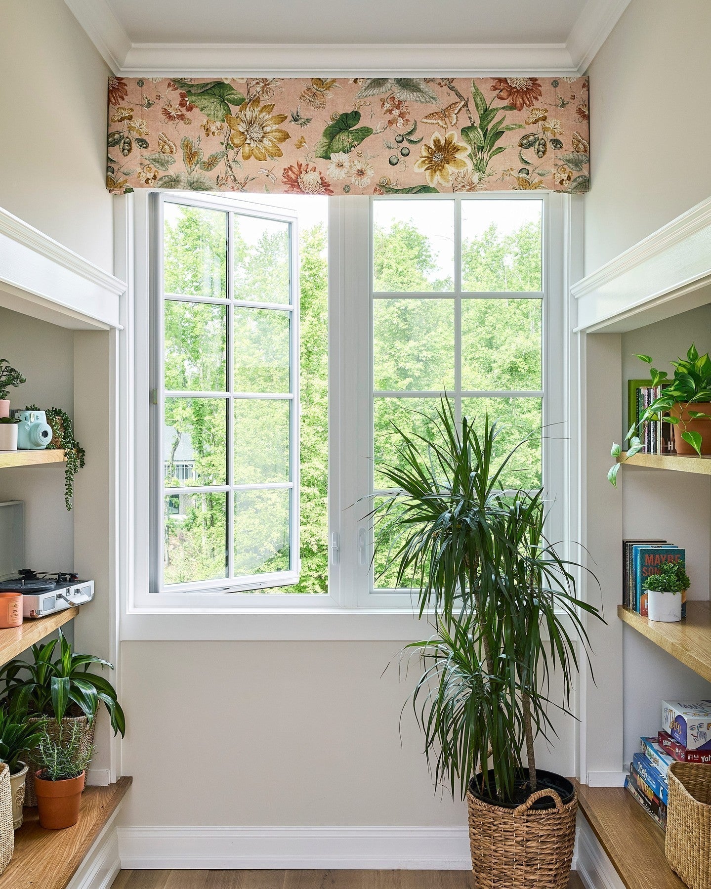 Patterned fabric Roman shades in flat fold style, light filtering and blackout options, embellishing a bright window space with plants and shelves.
