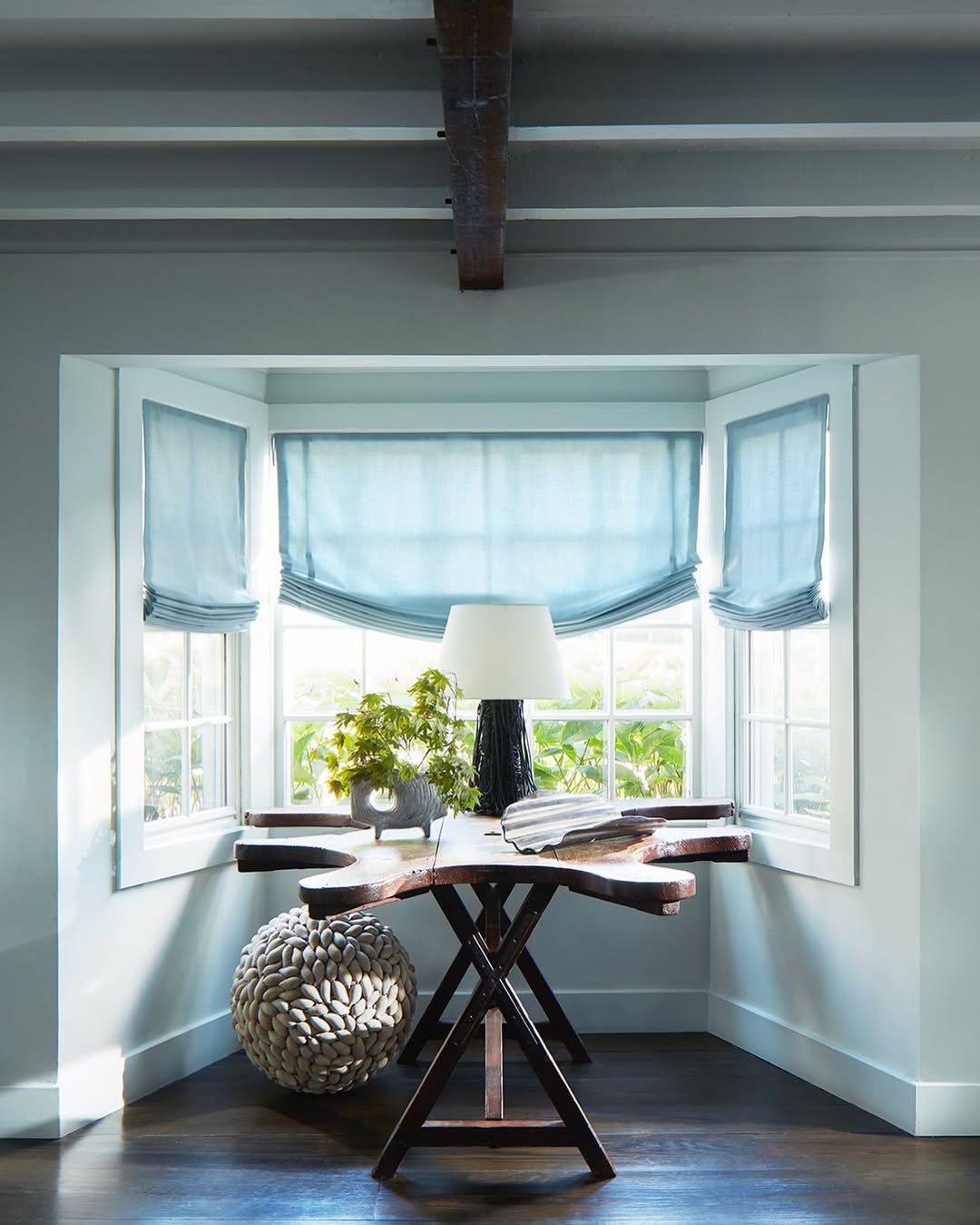 Light blue relaxed Roman shades with soft curved bottoms in a bright bay window setting with natural light filtering through