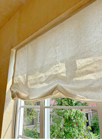 A beige linen Roman blind roller partially drawn over a white-framed window, with a warm yellow wall and green garden foliage visible outside.