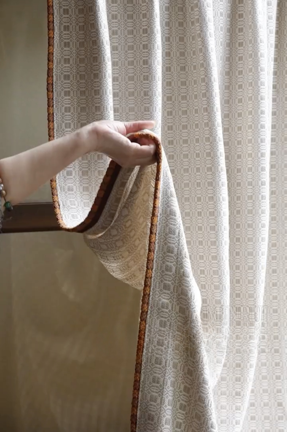 Hand holding edge of beige vintage curtains with geometric pattern and brown trim in soft natural light