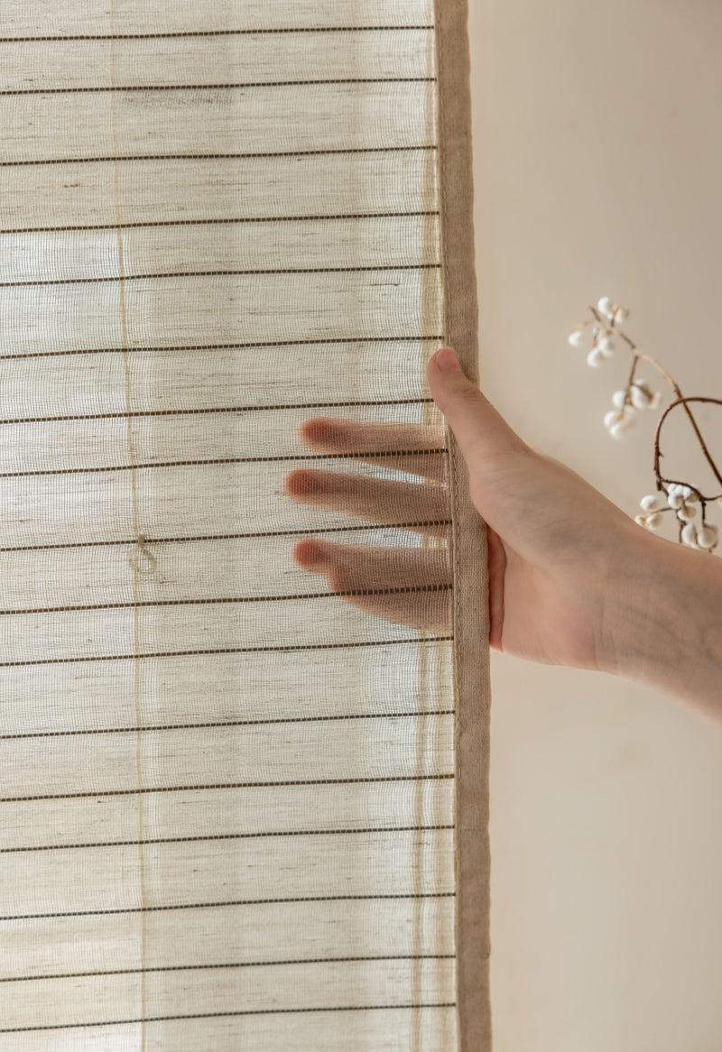 Hand holding a natural linen bamboo shade with black stripes, showcasing eco-friendly woven design and artisan craftsmanship.