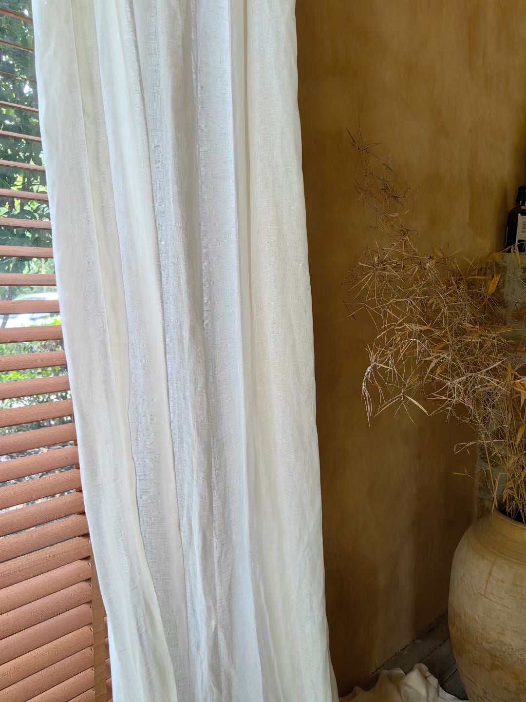 Pure white French-imported linen curtain hanging by a sunlit window, enhancing the serene decor of a room with a rustic yellow wall and natural elements