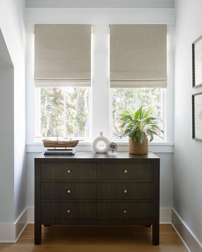 Eco-friendly beige Roman blinds in a stylish interior with wooden dresser and potted plant