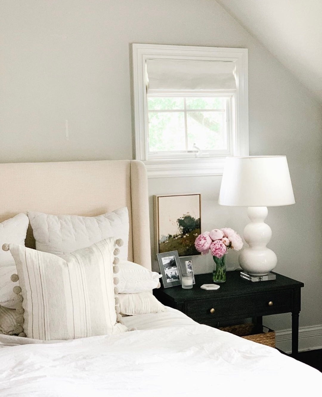 Elegant bedroom with beige sofa bed, plush pillows, black nightstand with white lamp and pink peonies, promoting a serene and stylish sleep environment with EaseEase blackout Roman blinds