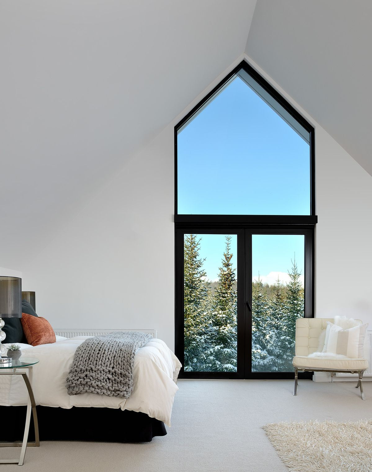 A modern bedroom with a large black-framed triangle window looking out onto snowy pine trees under a blue sky. The room has white walls, a bed with white bedding and gray throw, a cream armchair, and a light rug.