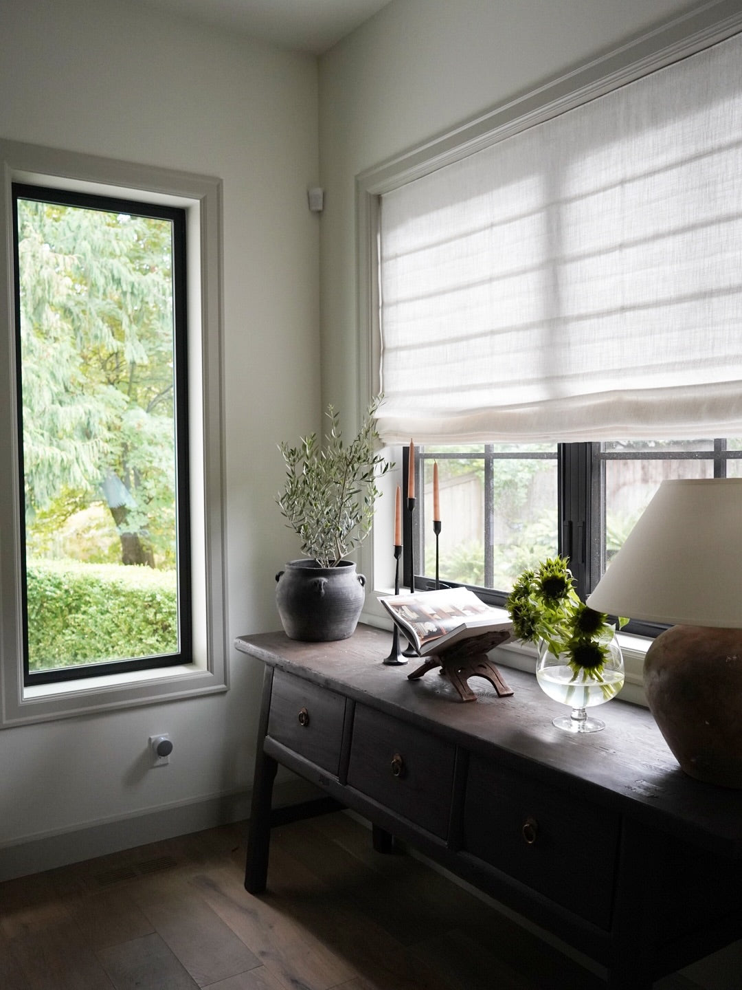 A dark wood console table in a sunlit room, styled with a potted olive branch, a small elephant figurine, a vase of greenery, and a table lamp. Two windows with white Roman shades and black frames overlook a green garden.