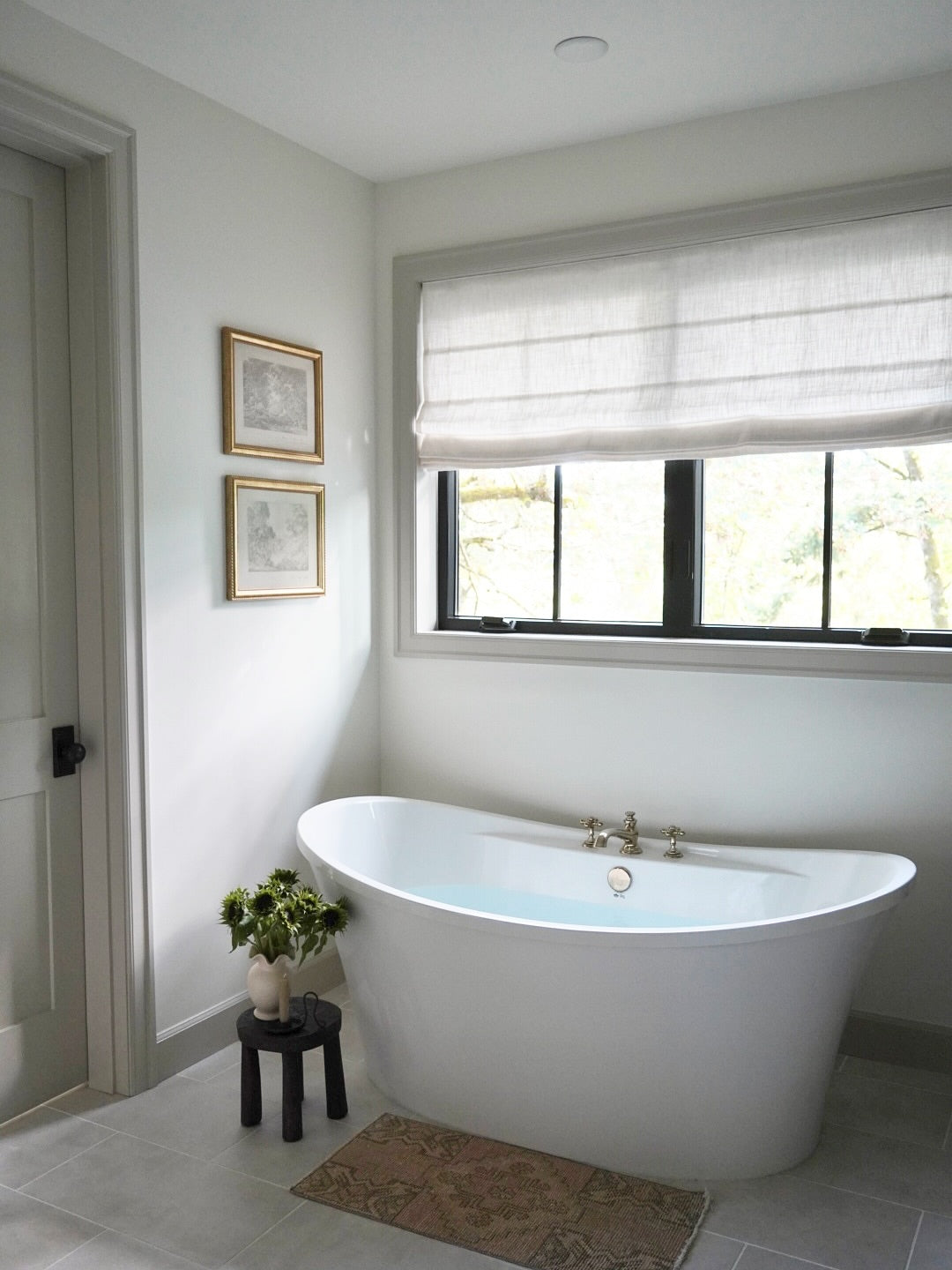 A white freestanding oval bathtub in a bright, modern bathroom with light gray walls, a window with a white roman shade, and a small black stool holding a potted plant beside the tub.