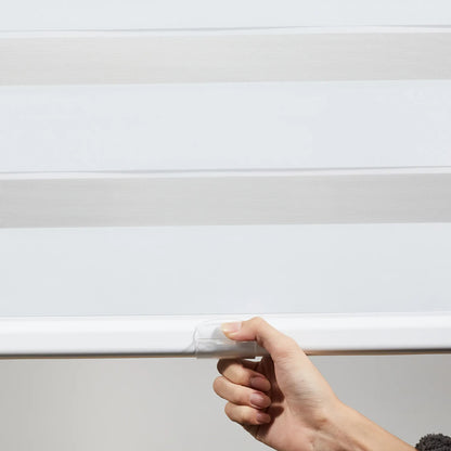 Close-up of a hand adjusting a white Zebra roller shade featuring alternating sheer and solid fabric bands, demonstrating the cordless system for customizable light control and privacy.