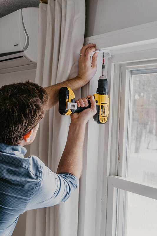 Professional curtain installation technician using a drill to install curtain hardware beside a window with white curtains.